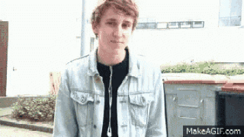 a young man wearing a denim jacket and earphones is standing in front of a trash can .