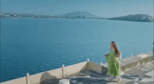 a woman in a green dress is standing on a dock overlooking a body of water .
