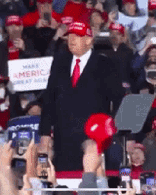 a man in a suit and tie stands in front of a crowd holding a sign that says " make america great again "
