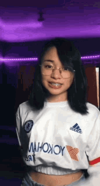 a woman wearing glasses and a white adidas shirt is standing in front of a purple background .