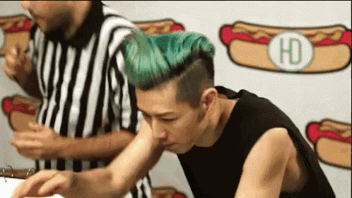 a man with green hair is eating a hot dog with a referee standing behind him .