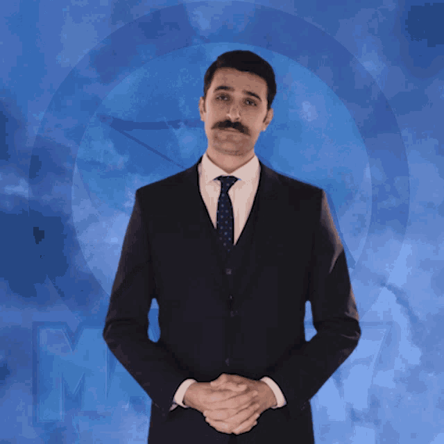 a man in a suit and tie is standing with his hands folded in front of a blue background with the letter m on it