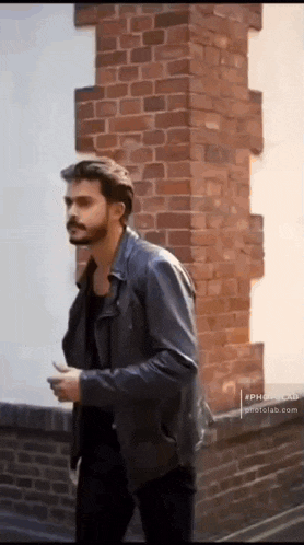 a man wearing a black leather jacket is walking in front of a brick wall .