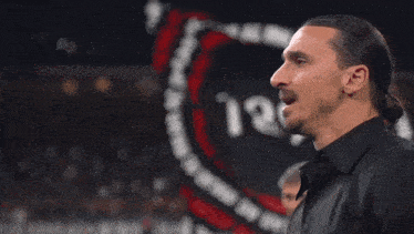 a man with a ponytail and a mustache is standing in front of a sign that says ' nm '