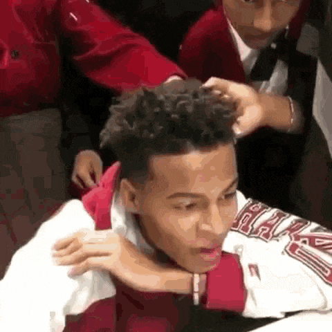 a man in a red jacket is getting his hair braided by another man .
