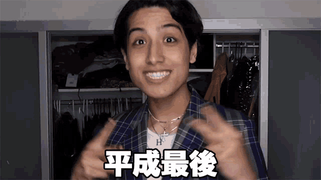 a man wearing a plaid jacket and a necklace with the letter h on it is standing in front of a closet
