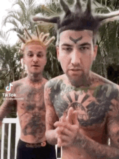 two men with tattoos and dreadlocks are standing next to each other . one of the men is wearing a crown on his head .