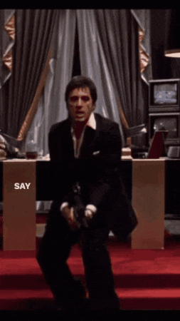 a man in a suit is sitting on a red carpet and holding a gun with the words say hello to my little friend above him