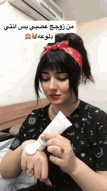 a woman wearing a bandana and a ring is holding a tube of lotion on her wrist