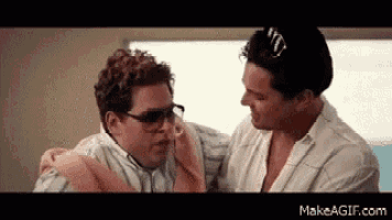 two men are sitting next to each other in a room . one of the men is wearing sunglasses .