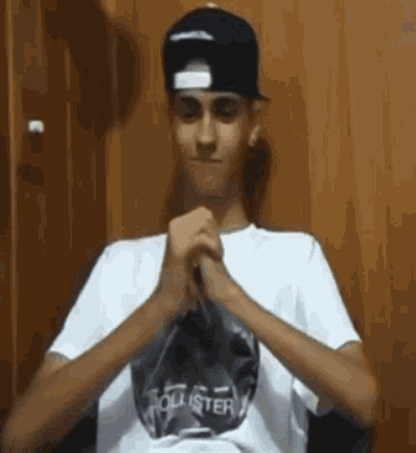 a young man wearing a hat and a hollister t-shirt is sitting in a chair with his hands folded .
