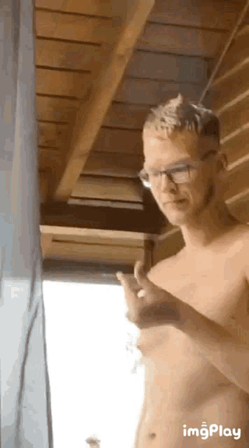 a shirtless man with glasses is standing in front of a window and giving the middle finger .