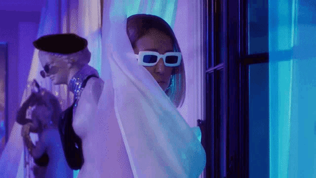 a woman wearing white sunglasses and a white scarf stands in front of a window
