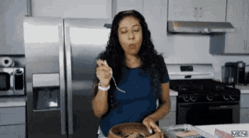 a woman in a blue shirt is eating food in a kitchen with a spoon .