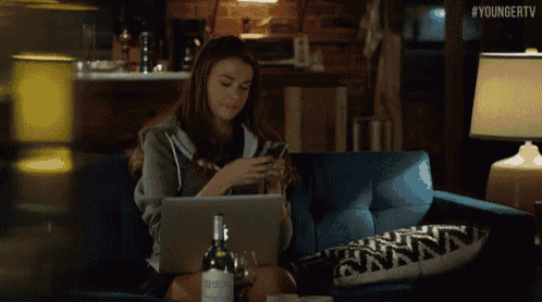 a woman sits on a couch with a laptop and a bottle of wine while looking at her phone