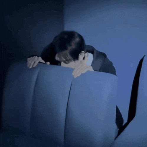 a man laying on a blue couch covering his face with his hands
