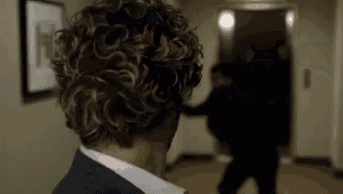 a man with curly hair is standing in a hallway looking at another man in a suit .