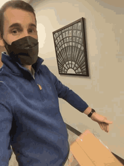 a man wearing a mask holds a cardboard box in front of a picture on the wall