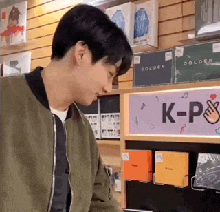 a man in a green jacket is standing in front of a shelf with k-pop albums .