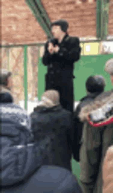 a man in a black coat is standing in front of a crowd of people and holding a microphone .
