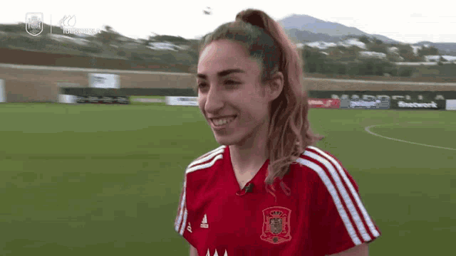 a woman in a red adidas jersey smiles on a field