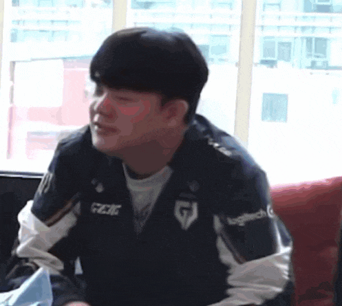 a man in a jacket is sitting on a couch in front of a window and making a funny face .