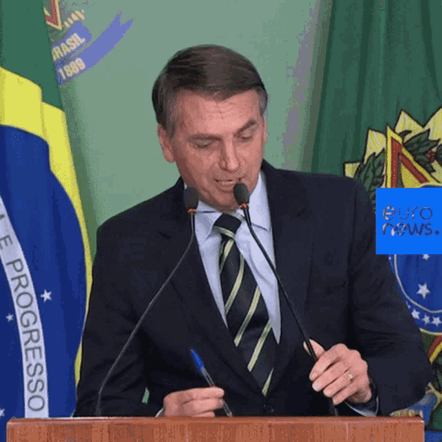 a man in a suit and tie stands at a podium holding a pen in front of a flag that says brasil 1889