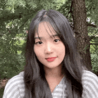 a young woman with long black hair and a striped shirt is standing in front of a tree .