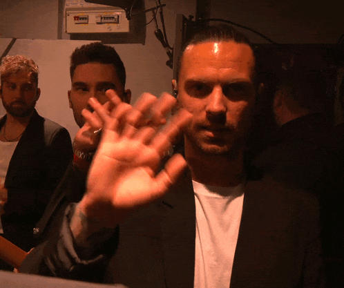 a man in a suit and white shirt waves his hand in front of a wall with a sign that says ' axx '