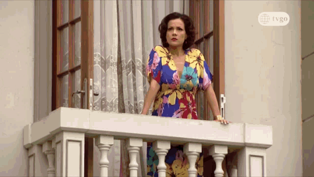 a woman in a floral dress stands on a balcony with a tvgo logo