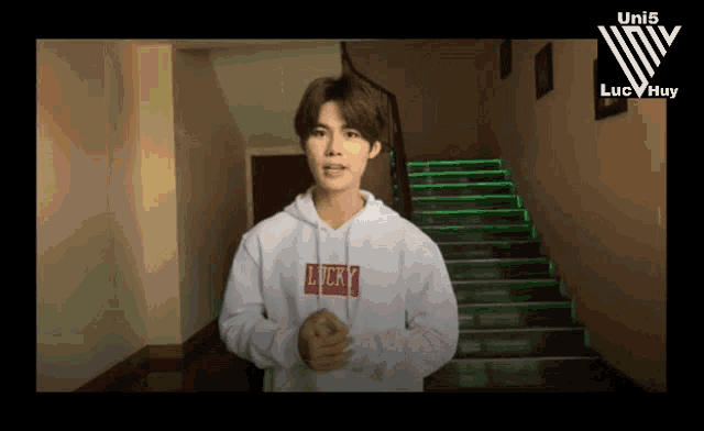 a young man wearing a white lucky sweatshirt stands in front of stairs
