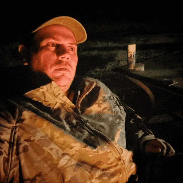 a man in a camouflage jacket sits in a dark room with a bottle of sk on the table