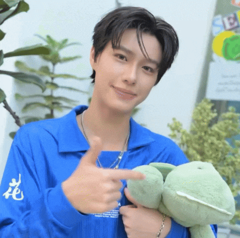 a young man in a blue shirt is holding a stuffed frog and pointing at it .