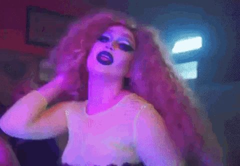 a drag queen with purple hair and black makeup is dancing in a club .