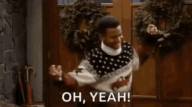 a man wearing a christmas sweater is standing in front of a door and saying `` oh , yeah ! ''