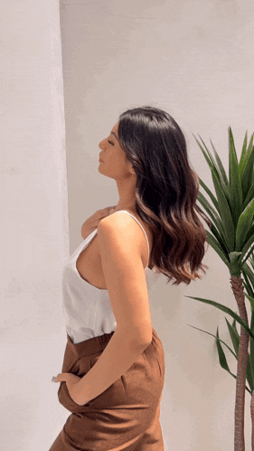 a woman in a white tank top and brown pants stands in front of a palm tree