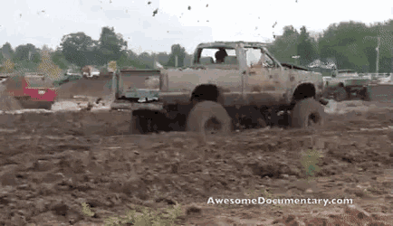 a truck is driving through a muddy field with a red truck in the background .