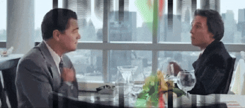 two men are sitting at a table in front of a window talking .