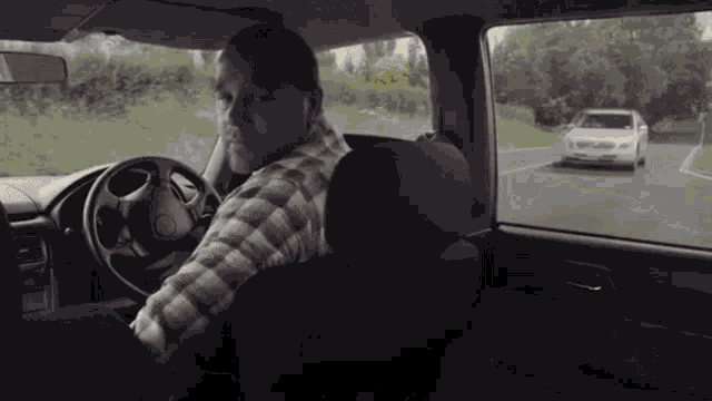 a man in a plaid shirt is driving a car down a road