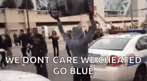 a group of people are standing in front of a police car and a man is holding a barrel over his head .