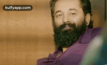 a man with a beard and mustache is wearing a purple shirt and looking at the camera .
