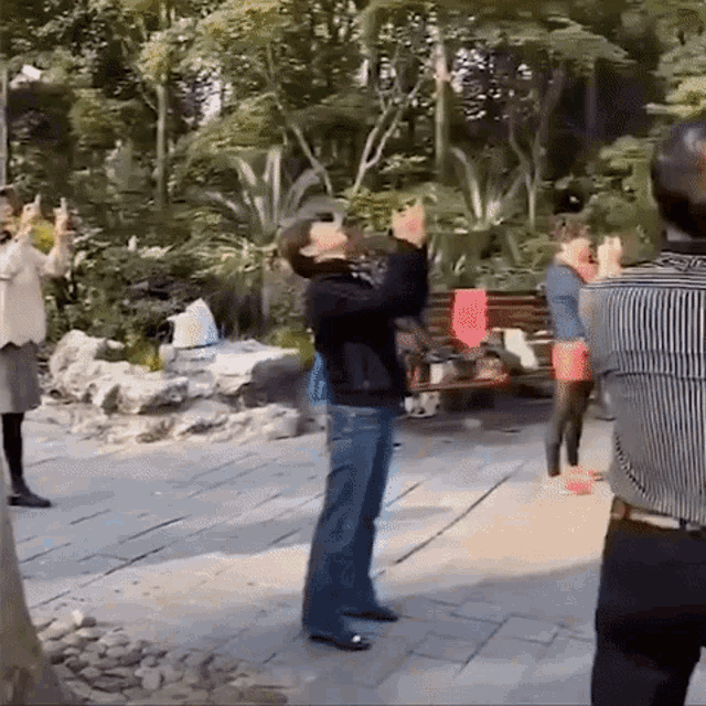 a group of people are standing on a sidewalk in a park taking pictures with their phones .