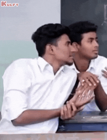 two young men are sitting at a desk in front of a blackboard .