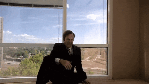 a man in a suit and tie sits on the floor in front of a large window