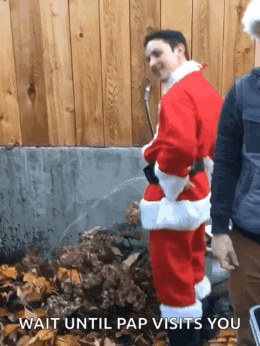 a man in a santa suit is standing in front of a wooden fence with the caption " wait until pap visits you "