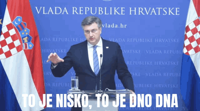 a man in a suit and tie stands at a podium in front of a vlada republike hrvatske banner