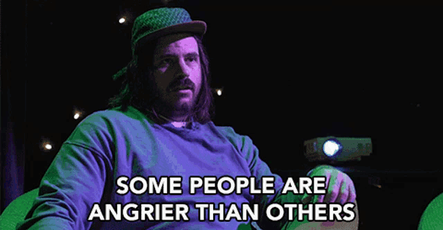 a man with long hair and a beard is sitting in front of a projector and says " some people are angrier than others "