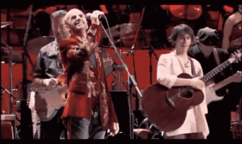 a man in a red jacket sings into a microphone while a woman plays a guitar