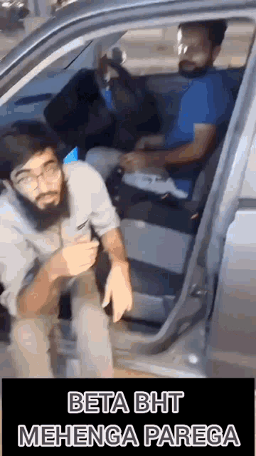 two men are sitting in a car with the door open and one of them is wearing glasses .