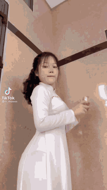 a girl in a white dress is standing in front of a wall and holding a bottle of perfume .